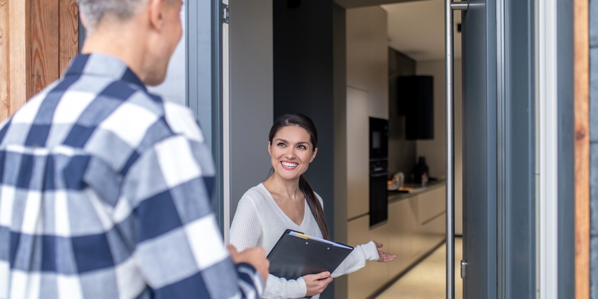 Atendente sorrindo recebe um cliente na porta de um imóvel, com uma prancheta na mão